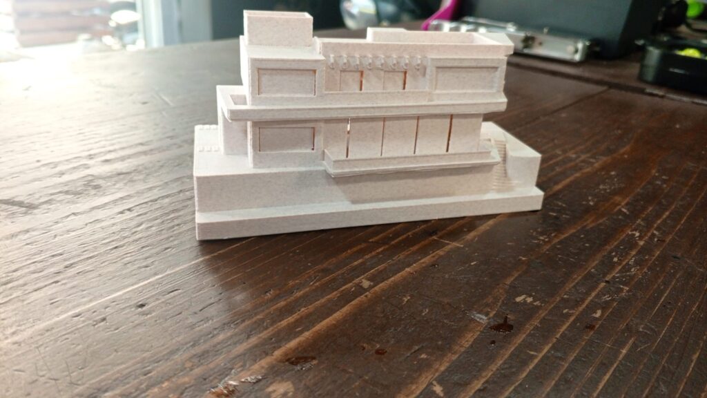 Small white building model with a stepped base and projecting roof.